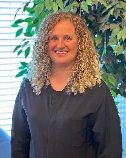A woman with curly hair smiles while standing indoors against a leafy background. She is dressed in dark professional clothing.