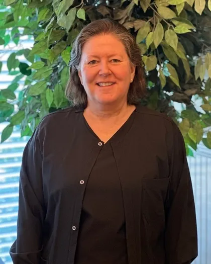 A woman smiles while standing indoors in front of leafy greenery. She is wearing dark professional attire.