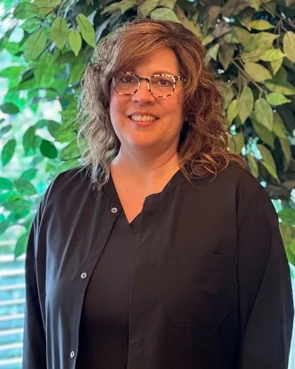 A woman wearing glasses smiles while standing indoors against a leafy background. She is dressed in dark professional clothing.