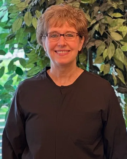 A woman with short hair and glasses smiles while standing in front of indoor greenery. She is dressed in dark professional attire.
