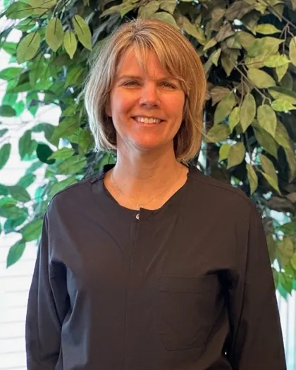 A woman with a blonde short hair, smiles while standing indoors in front of a green leafy backdrop. She is wearing dark professional clothing.