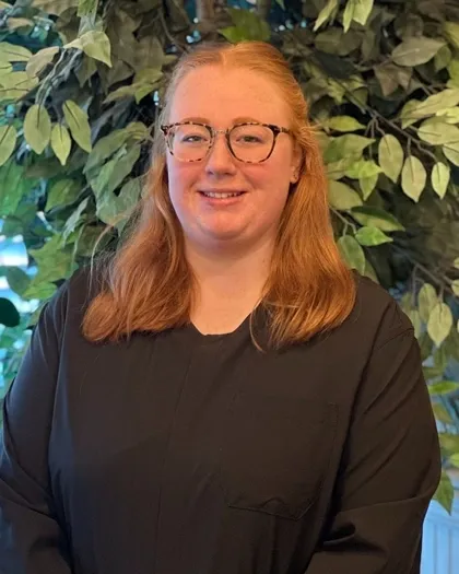 A woman wearing glasses smiles while posing indoors with leafy plants behind her. She is dressed in dark professional attire.