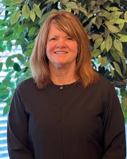 A woman with shoulder-length hair smiles while standing in front of indoor greenery. She is dressed in dark professional clothing.