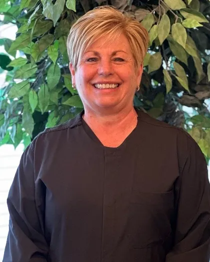 A woman smiles at the camera while standing indoors against a leafy background. She is wearing a dark professional top.