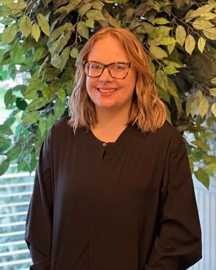 A woman wearing glasses smiles while standing indoors in front of leafy greenery. She is dressed in dark professional attire.