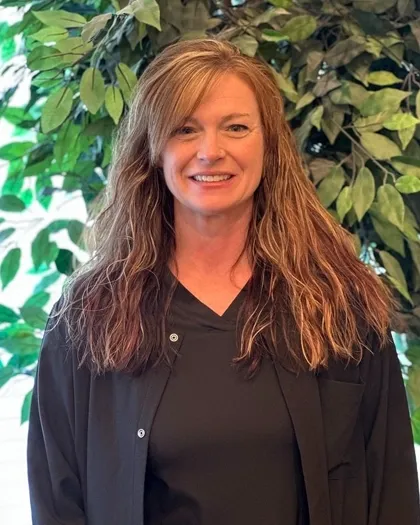 A woman with long hair smiles while standing in front of greenery indoors. She is dressed in dark professional clothing.