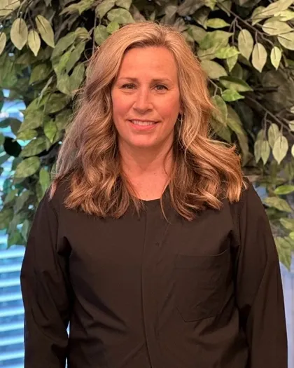 A woman stands smiling indoors against a leafy green background. She is wearing dark professional attire in a clinic or office setting.