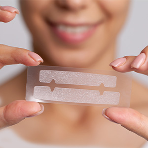 A smiling person holds a clear dental aligner in front of their mouth. The image highlights orthodontic treatment using clear aligners for teeth straightening.