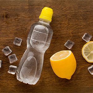 A bottle of lemon-flavored drink is placed on a wooden surface alongside lemon slices and ice. The image suggests acidic beverages that can impact dental enamel.