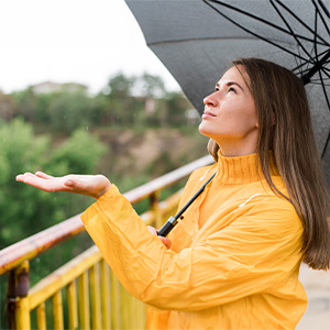 A woman stands outdoors holding an umbrella and looking upward as rain falls. The image conveys a lifestyle or environmental scene often used for wellness or mood-related context.