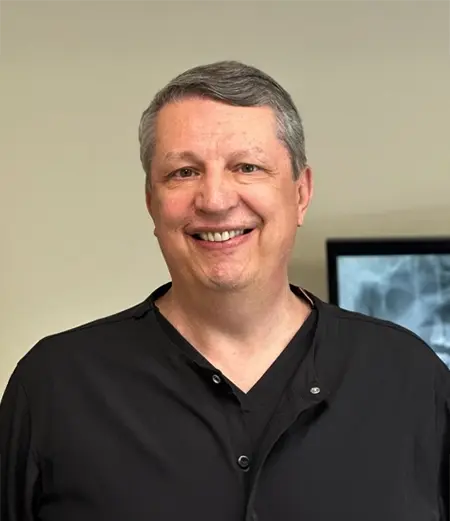 A man smiles while posing for a professional indoor portrait. He is wearing a dark shirt against a neutral background.