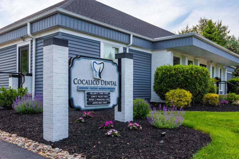 An exterior view of a dental office building shows a sign reading “Cocalico Dental” positioned in front of the property. The building features gray siding, white brick columns, and landscaped greenery surrounding the entrance.