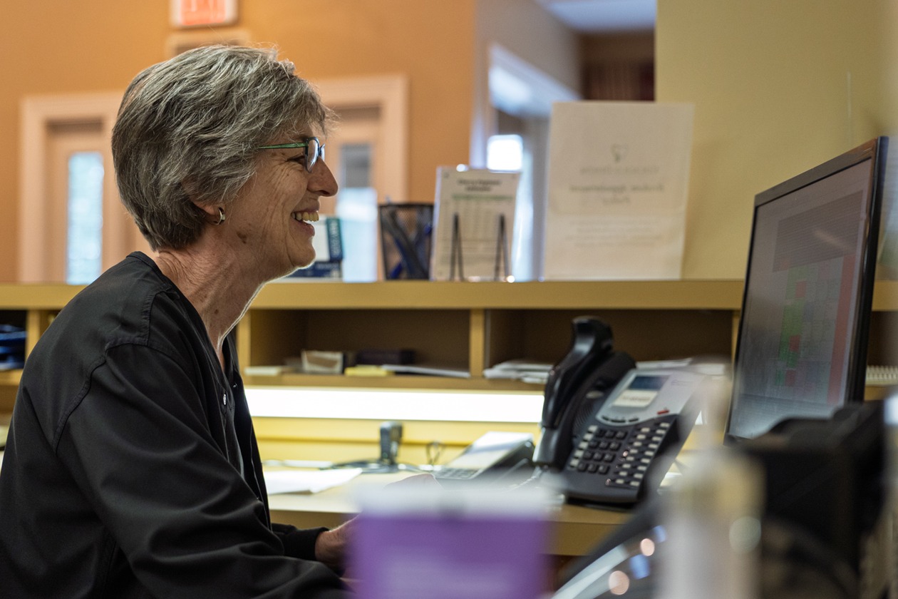 A woman smiling and sits alone at a dental front desk inside a clinic. Office equipment and paperwork are visible around the reception area.
