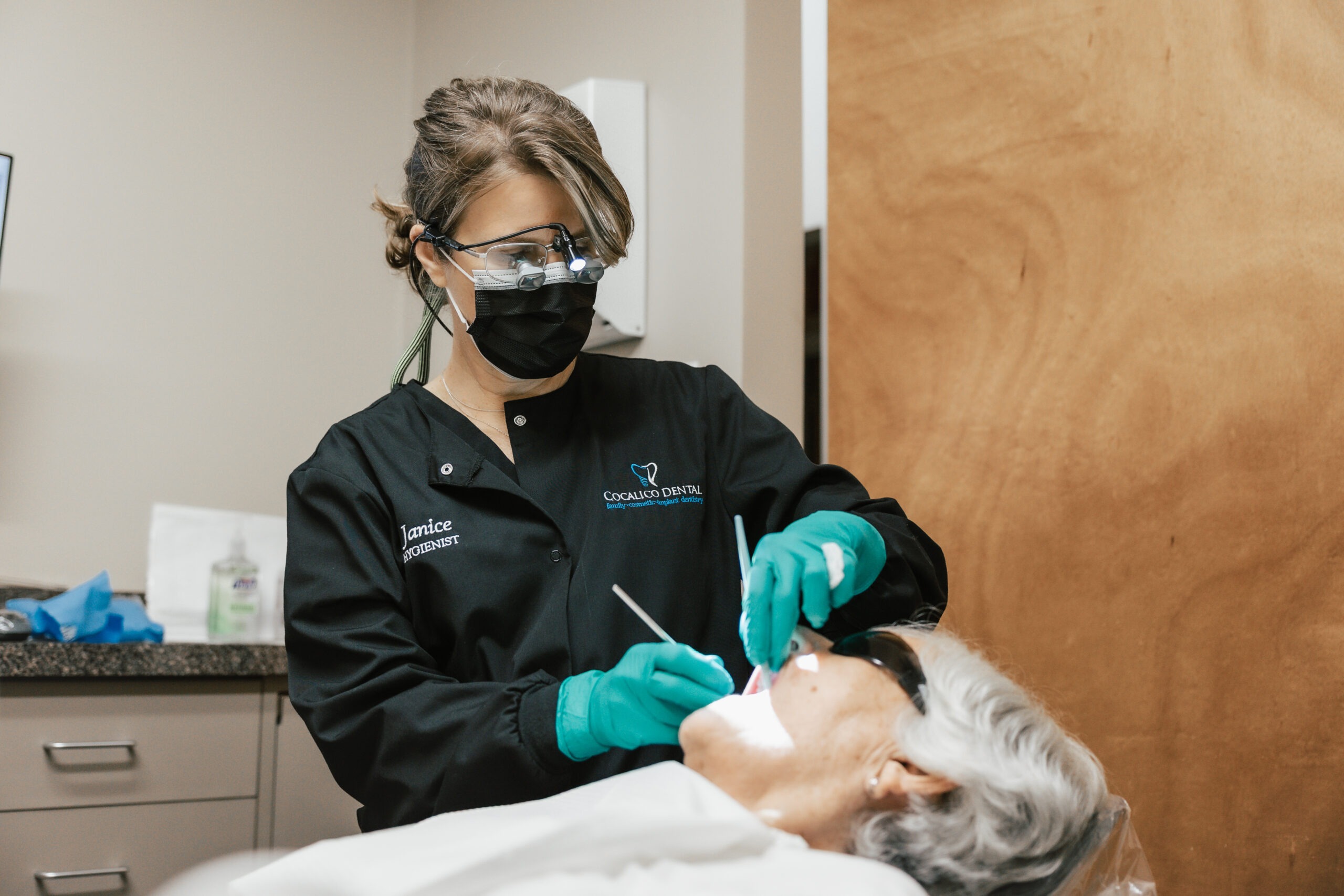 A dental professional treats a patient who is reclined in a dental chair. Gloves, instruments, and clinical equipment are visible during the procedure.